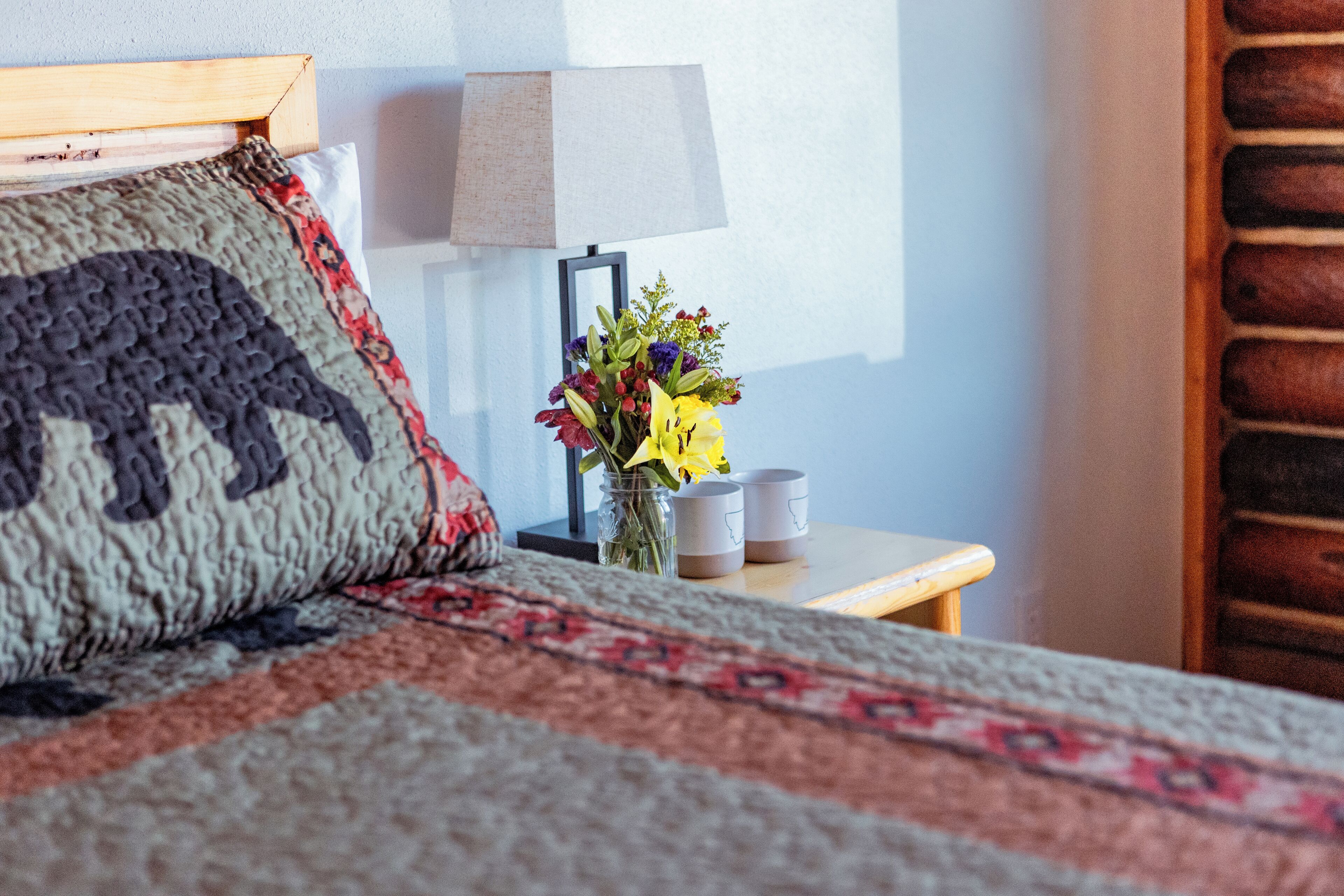 A neatly made bed with a patterned pillow, a bedside table with a lamp, and a vase with flowers.