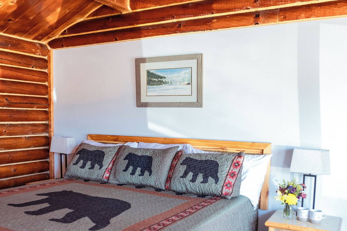 Teto de madeira, uma cama com manta temática de urso, uma mesinha de cabeceira com abajur e um quadro na parede.