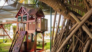 Una casetta gioco in legno con uno scivolo e diverse finestre, immersa in un ambiente naturale con alberi e un tavolo da picnic.