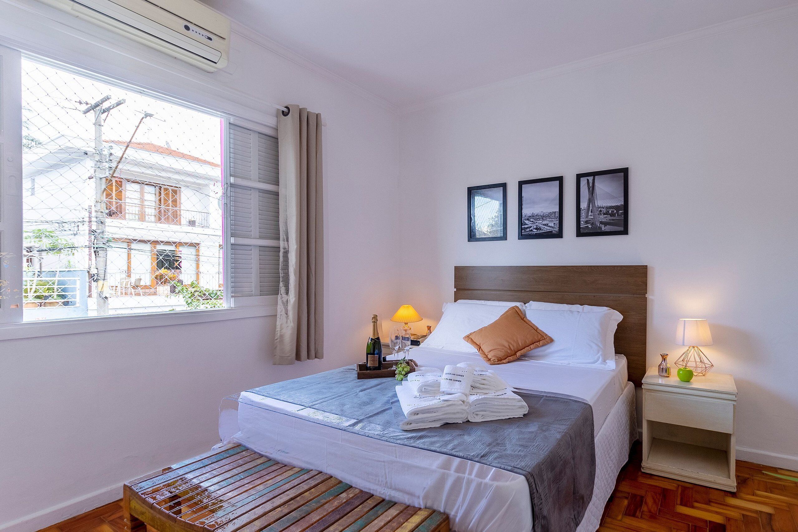 A neatly made bed with a blue bedspread, a wooden bench, a bedside table with a lamp, and three framed pictures on the wall.