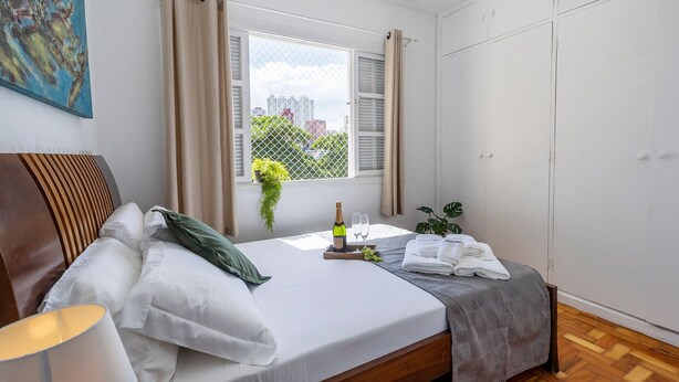 A neatly made bed with white linens, a bedside table with a lamp, a window with curtains, and a city view. - Casa com 4 quartos na Vila Madalena (São Paulo)