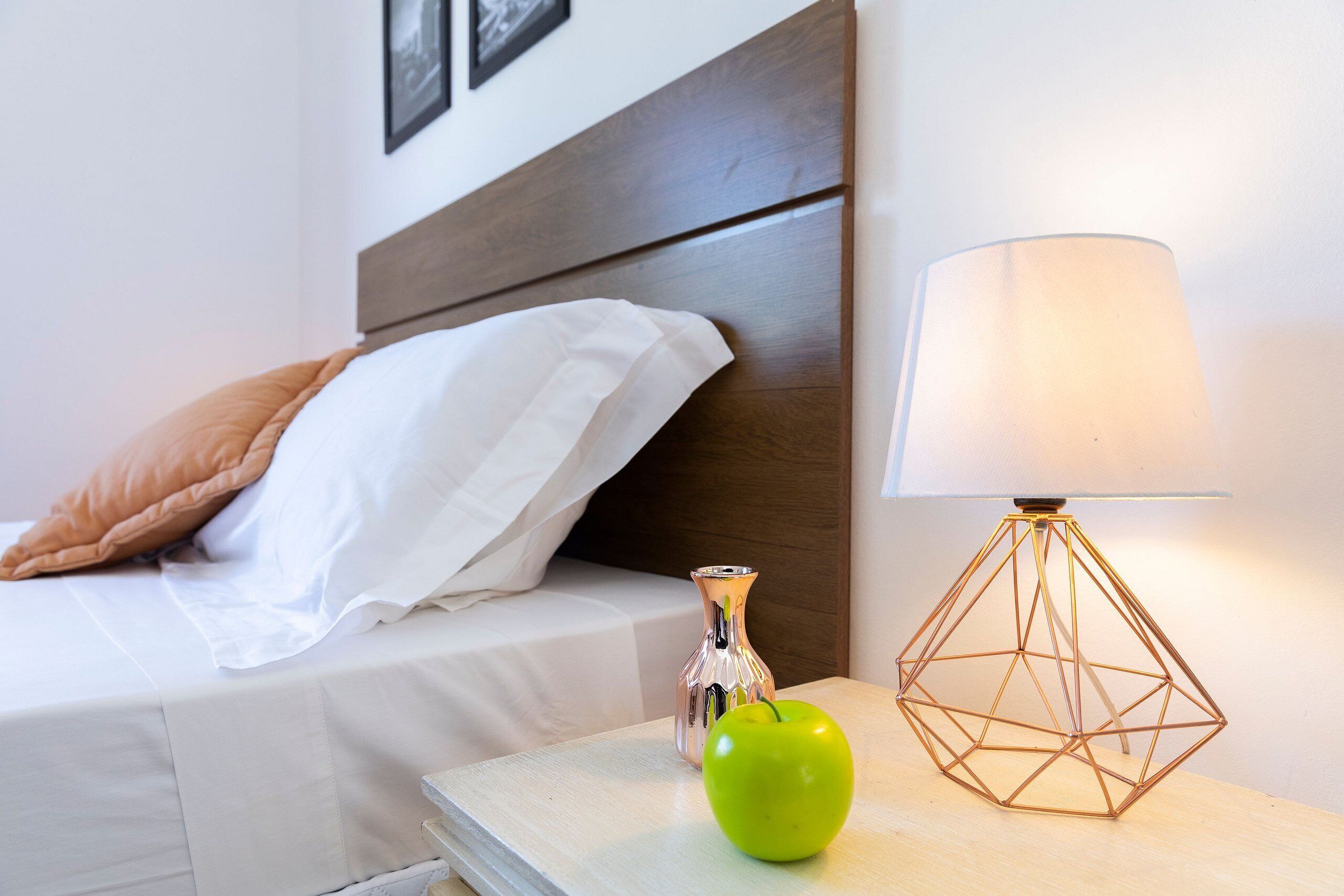A neatly made bed with a white comforter and a brown pillow, a clear glass vase, and a green apple on a bedside table next to a geometric lamp.