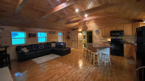 Een cabin interieur met een houten plafond, een zwarte leren bank, een keukenhoek met een magnetron en een koelkast, en een bordje met de tekst 'Nittany Lions'.