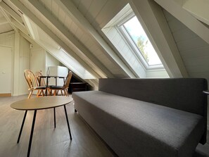 Une pièce de style « loft », avec un plafond haut, une lucarne, un canapé gris, une table ronde en bois et deux chaises en bois.