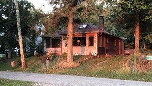 Une cabane en bois rustique, dotée d’une cheminée, entourée d’arbres et bordée d’une zone herbeuse.