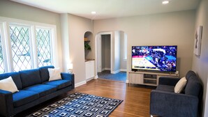 A living room with a blue sofa, a patterned rug, a TV displaying a sports event, and a hallway leading to another room. - Beautifully restored home in Butler Tarkington Walking Distance to Butler Campus (Indianapolis)