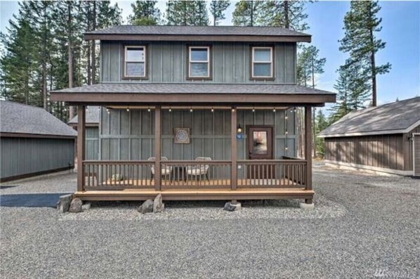 A two-story wooden cabin with a balcony, surrounded by gravel and trees.