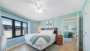 Une chambre à coucher comprenant un lit, un ventilateur de plafond, une fenêtre, une table de chevet, une lampe, un tapis et une porte donnant sur une autre pièce.