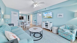 Un salon avec un canapé de couleur turquoise, une table basse ronde et un fauteuil à motifs. On y trouve également un coin repas avec une table et des chaises blanches, et une cuisine à l’arrière-plan.