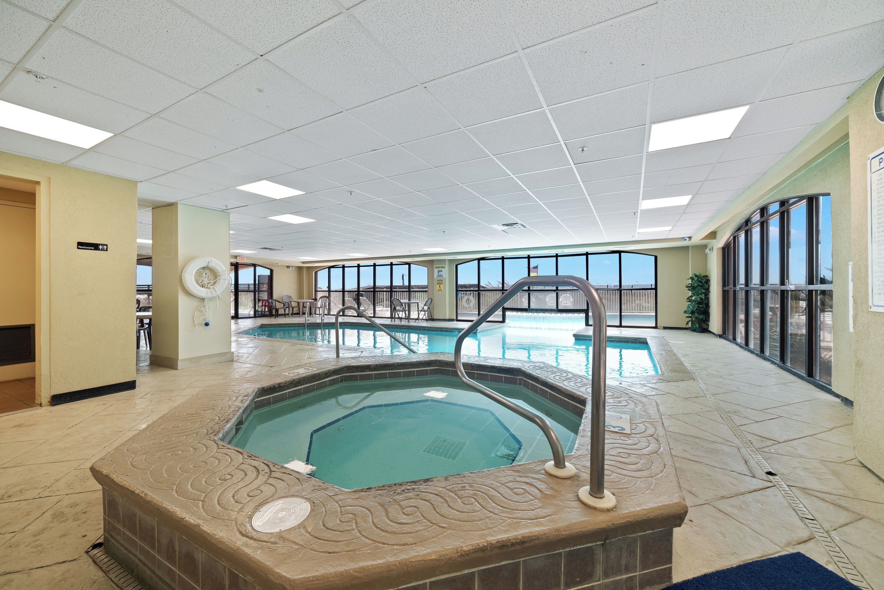 A large indoor swimming pool with a hot tub, surrounded by a tiled floor and large windows.