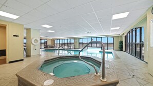 Une grande piscine intérieure avec un jacuzzi, entourée d’un sol carrelé et de grandes fenêtres.