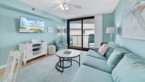 Un salon avec un canapé de couleur turquoise, un meuble de rangement blanc pour les appareils électroniques, un ventilateur de plafond et une grande fenêtre donnant sur l’extérieur.