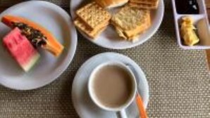 Une table mise pour le petit-déjeuner avec des assiettes de toasts, de la confiture et un bol de céréales. On y trouve également deux tasses de café et une assiette avec une tranche de pastèque et une tranche de pain grillé tartinée de confiture.