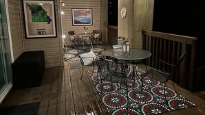 Een terras buiten met een houten dek, een metalen tafel en stoelen, en een vloerkleed met een patroon.
