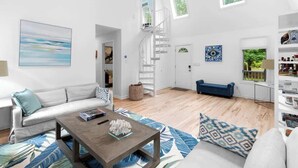 Een ruime woonkamer met een witte bank, een salontafel en een blauw tapijt met patroon. Er is een trap die naar een hoger niveau leidt, en een boekenkast vol boeken en decoratieve objecten.