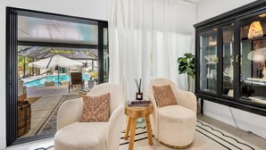 A modern living room with a pool view, two plush armchairs, a small wooden side table, and a black cabinet with glass doors. - Spacious Family Retreat with Pool Gym Sauna Pickleball and Guest Suite SLEEPS 16 (Naples)