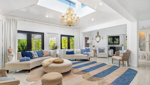 A spacious living room with a large sofa set, ottomans, and a patterned rug. There is a chandelier, a television, and a kitchen area visible. - Spacious Family Retreat with Pool Gym Sauna Pickleball and Guest Suite SLEEPS 16 (Naples)