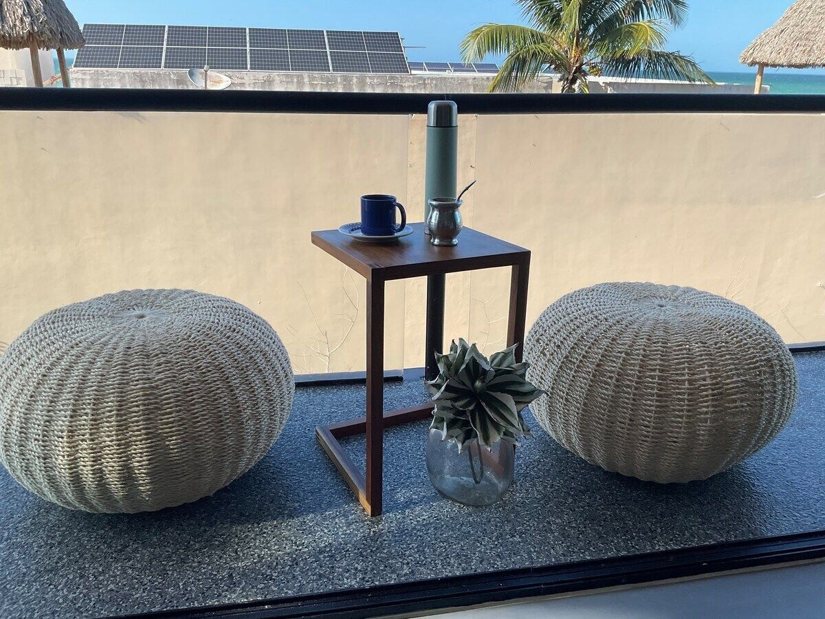 A small table with a blue cup and a thermos, flanked by two large woven poufs, with a view of solar panels and palm trees in the background.