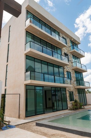 A modern multi-story building with large glass windows and balconies, a stone accent wall, and a swimming pool in the foreground.