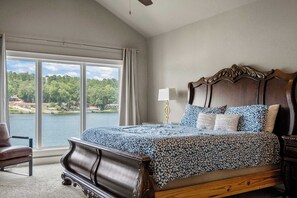 A bedroom with a large window, a bed with patterned bedding, a wooden headboard, and a nightstand with a lamp. - Luxe Waterfront Home in Hot Springs (Hot Springs)
