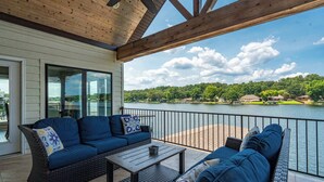 En overdækket terrasse med fletningsmøbler, et bord og udsigt til sø og skov.