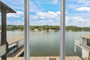 A view through a window of a lake with a wooden deck, a white railing, and a building across the water. - Luxe Waterfront Home in Hot Springs (Hot Springs)