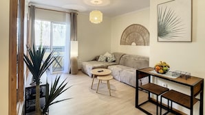 Ein modernes Wohnzimmer mit einer Couch, einem runden Tisch und einem Couchtisch. Eine große Pflanze, eine Lampe und ein an der Wand befestigtes Kunstwerk sind ebenfalls vorhanden.