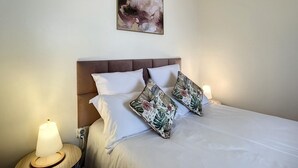 Una cama bien tendida con sábanas blancas y dos almohadas estampadas con motivos florales, un cabecero de madera y un cuadro abstracto colgado en la pared.