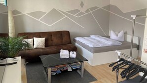 A modern hotel room with a brown sofa, a glass coffee table with magazines, a bed with white linens, and a clothing rack.