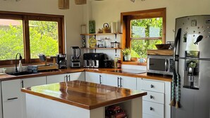 Una cocina con vigas de madera, una isla central, armarios blancos, un refrigerador y una ventana con vistas a la vegetación.