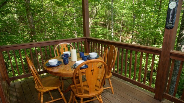 A wooden deck with a table and chairs, surrounded by a wooden railing, overlooking a forested area.