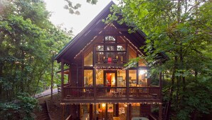 A two-story wooden cabin with a balcony, surrounded by trees.