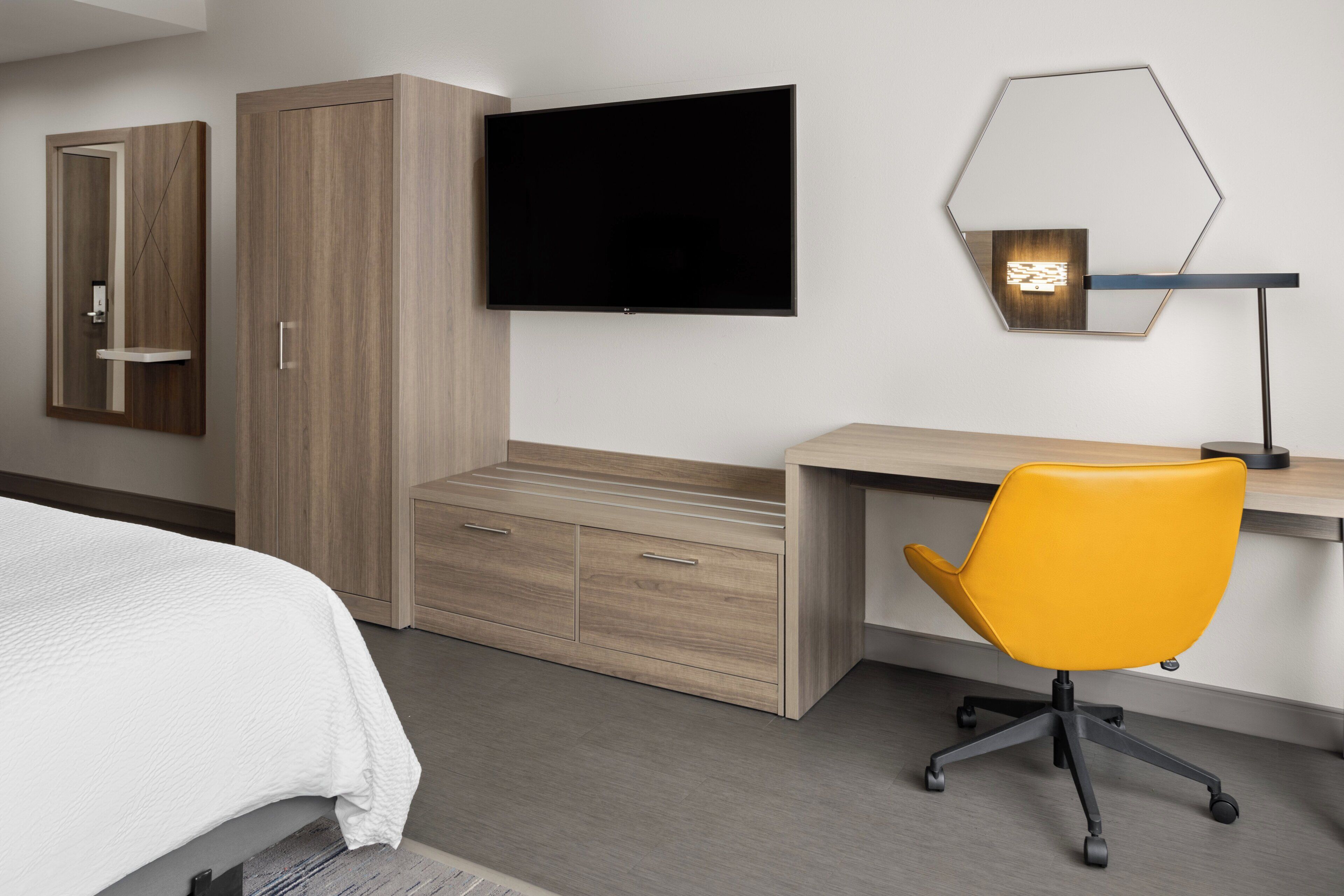 A hotel room with a bed, a wooden wardrobe, a flat-screen TV, a desk with a yellow office chair, and a unique hexagonal mirror.