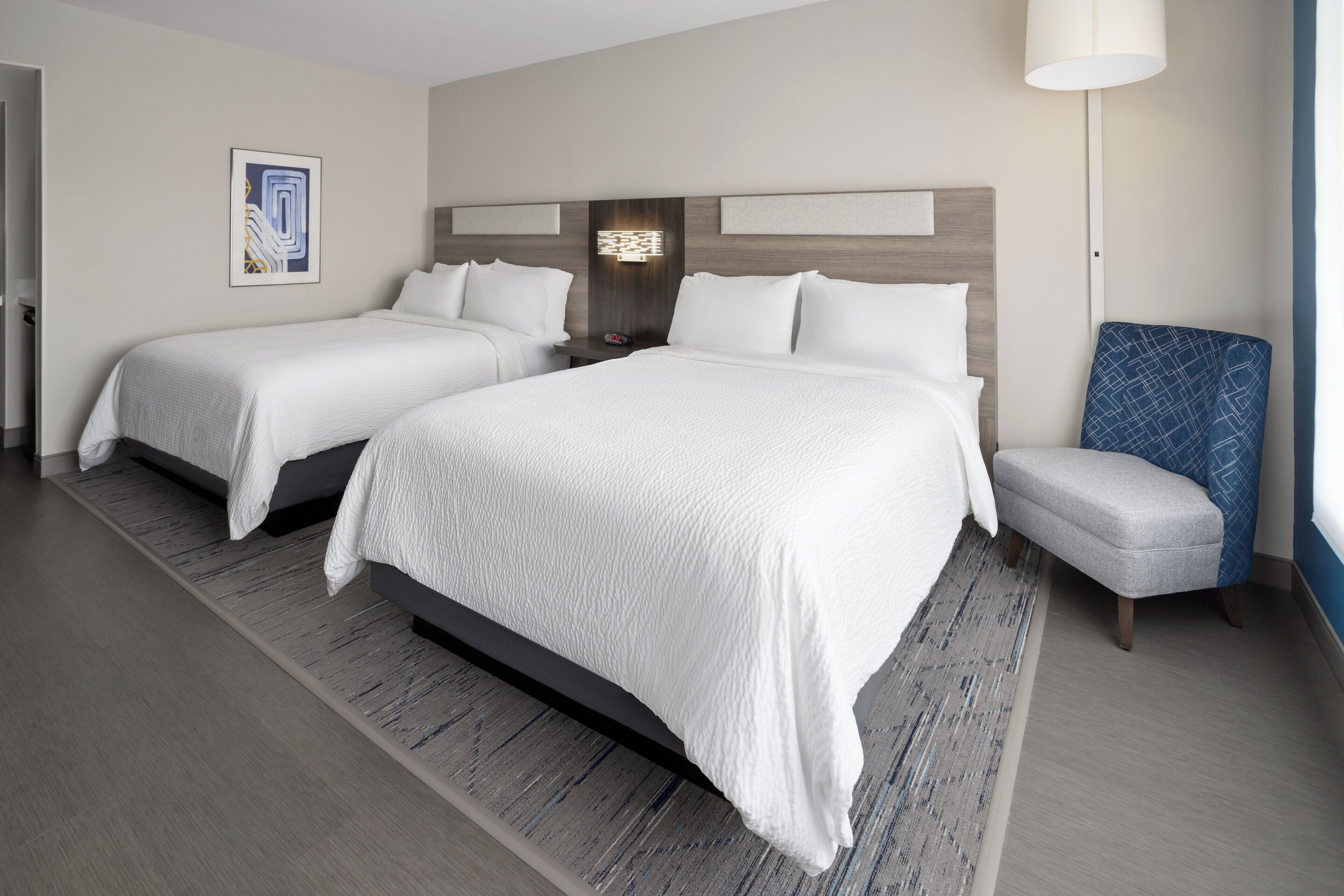 A hotel room with two beds, a nightstand, a lamp, and a chair.