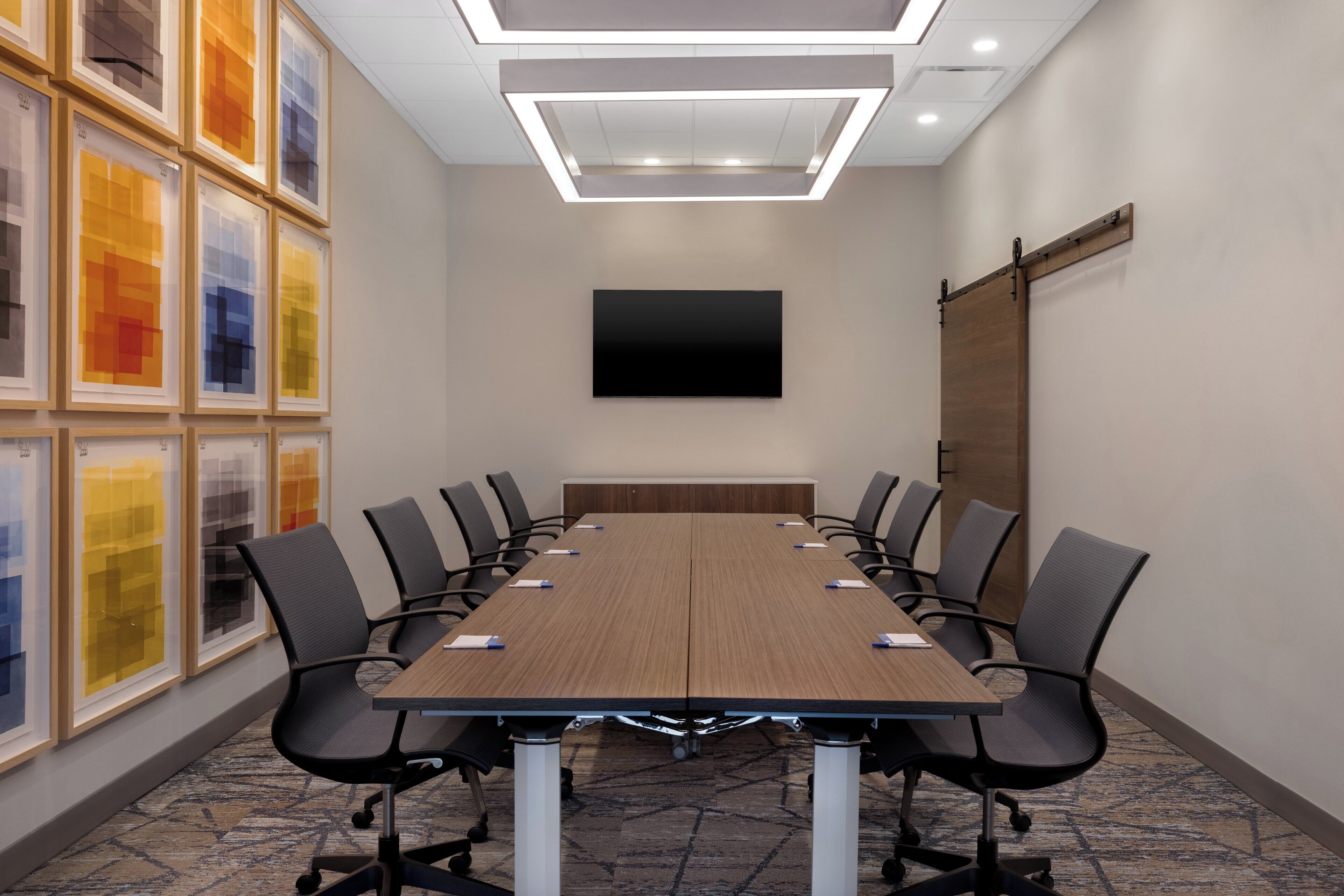 A conference room with a long table, chairs, a flat-screen TV, and framed artwork on the wall.