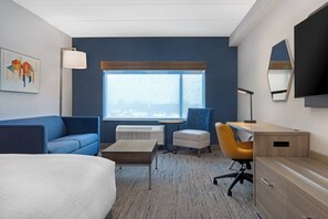 A modern hotel room with a bed, a desk, a chair, a sofa, a small table, a lamp, a window with a view, and a mirror.