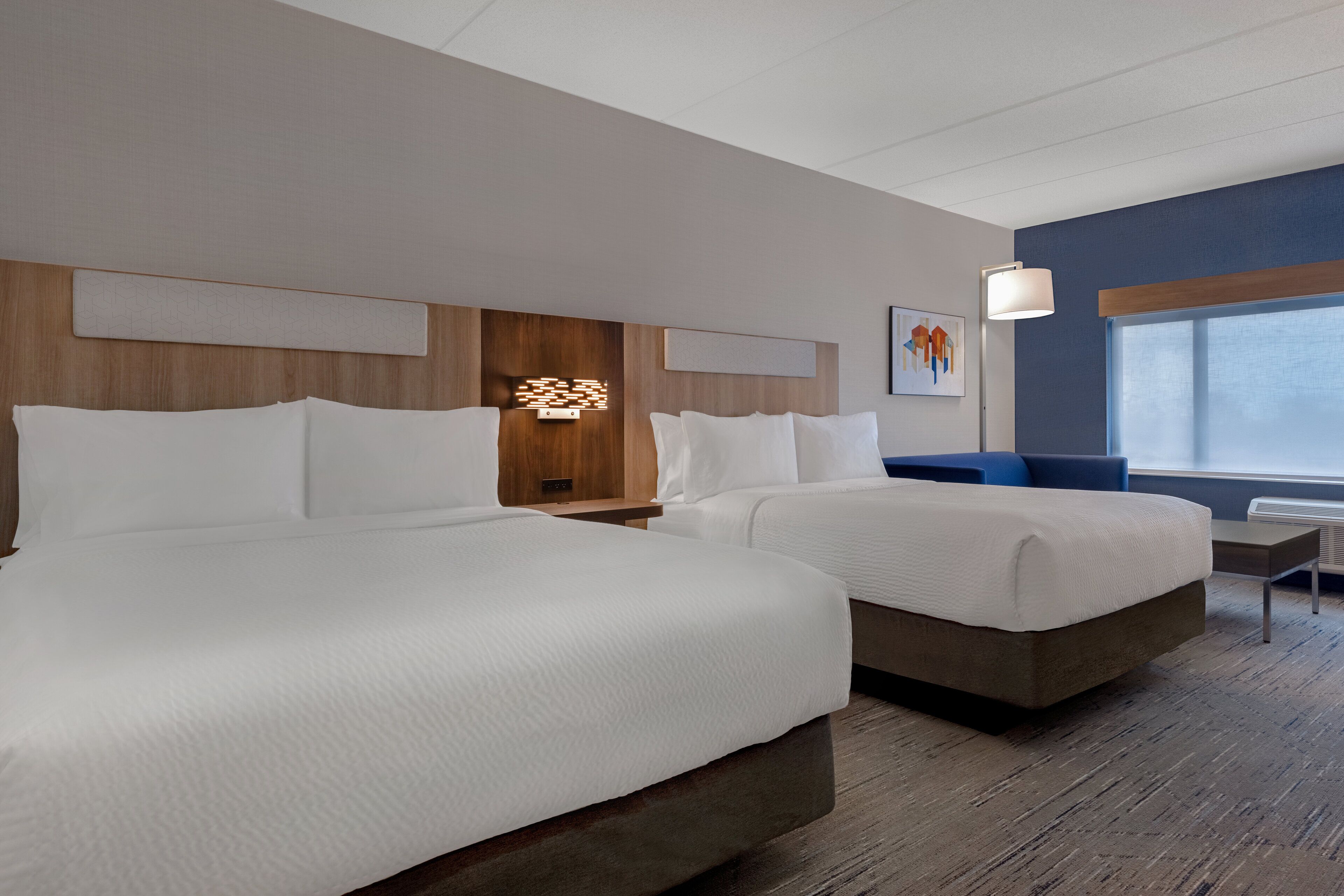 A hotel room with two beds, a wooden headboard, a wall-mounted light, and a window with a view.