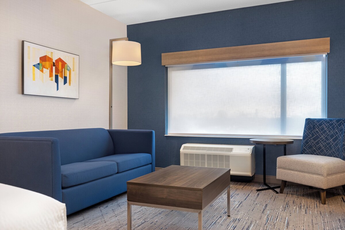 Une chambre d’hôtel moderne avec un canapé bleu, une table basse en bois, une petite table ronde, un fauteuil beige, une grande fenêtre avec des stores, un radiateur au sol et une lampe fixée au mur.
