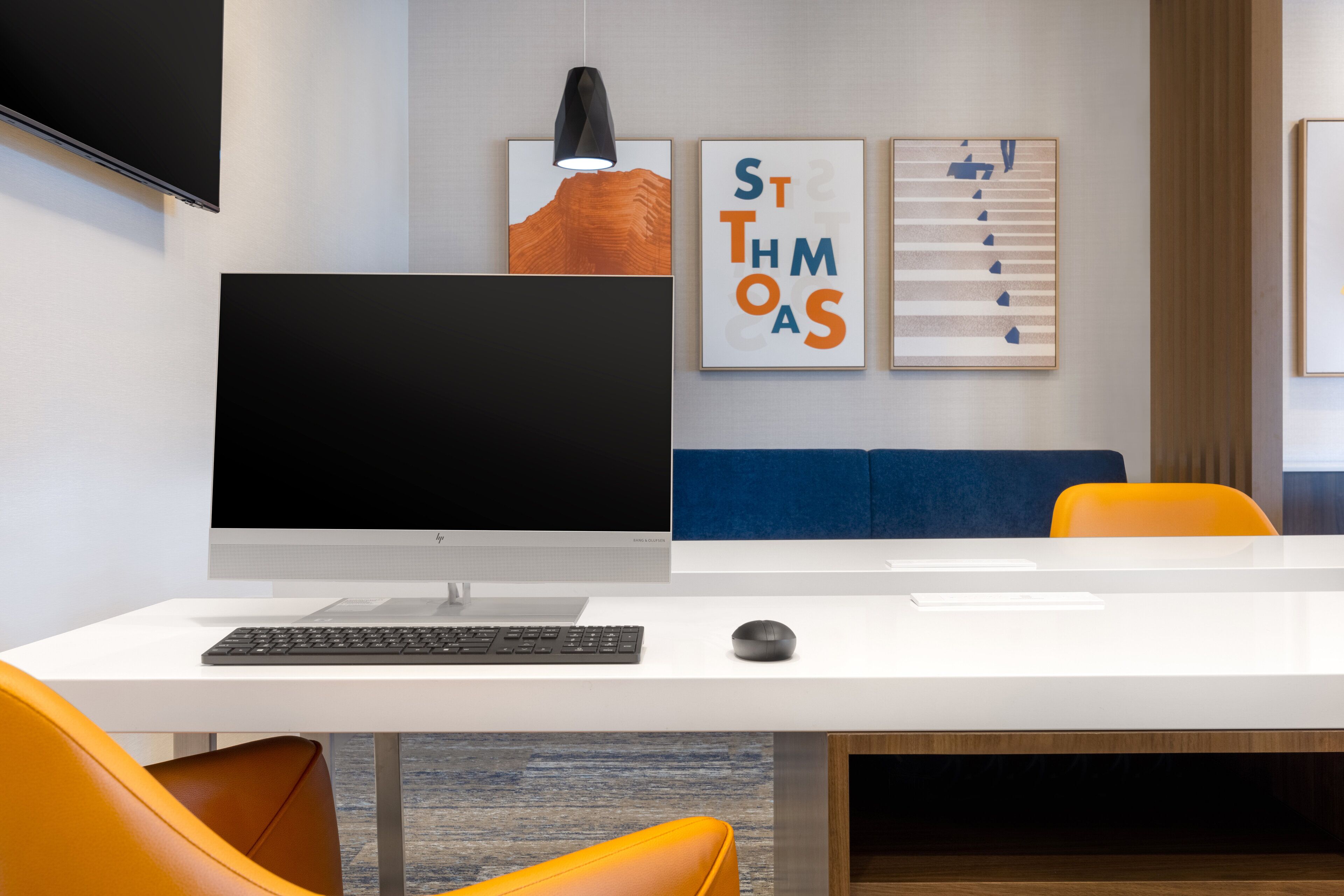 A modern office space with a white desk, a computer monitor, a keyboard, and a mouse. There are two framed artworks on the wall, a sofa, and an orange chair.