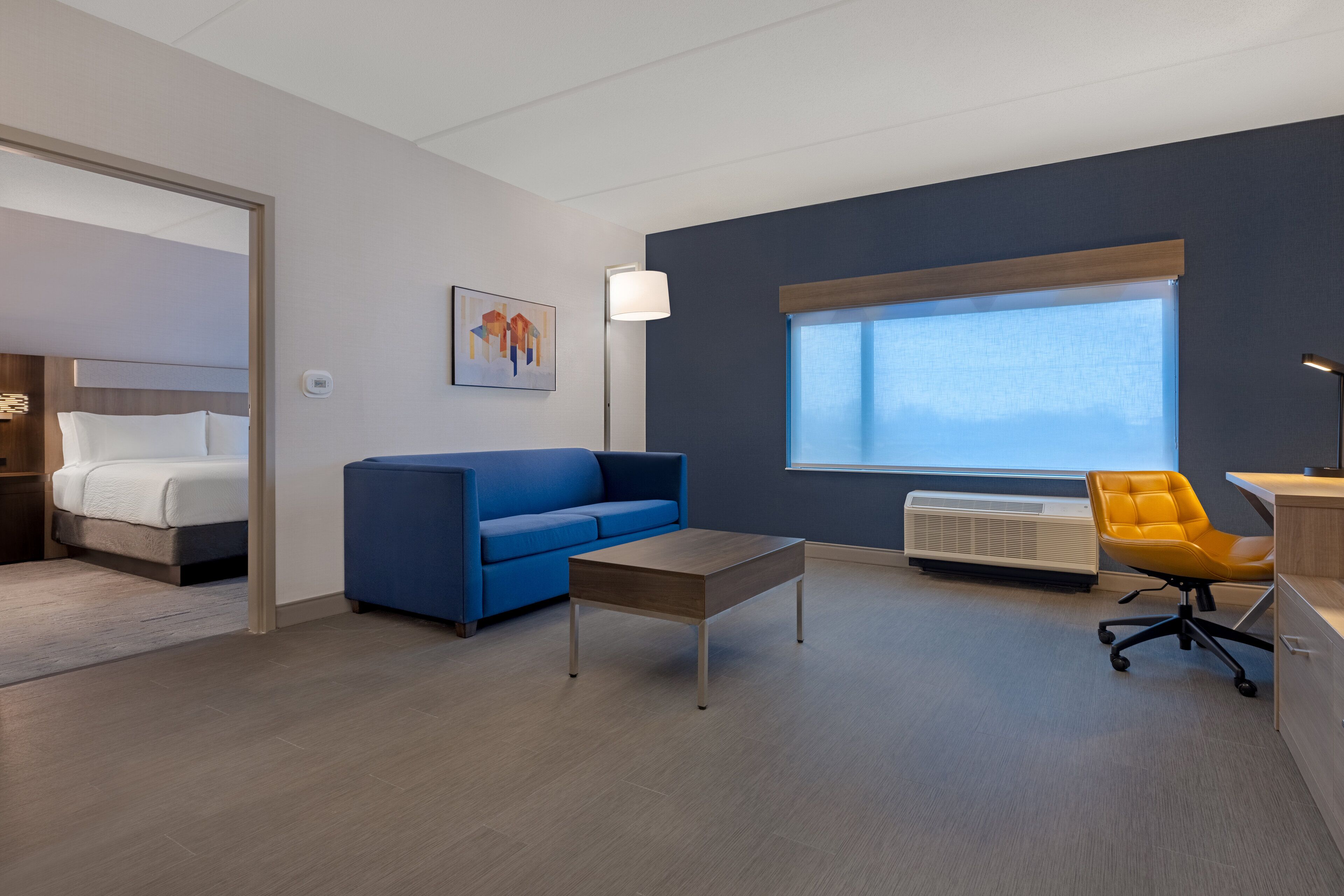 A modern hotel room with a bed, blue sofa, wooden coffee table, and a desk with a chair.