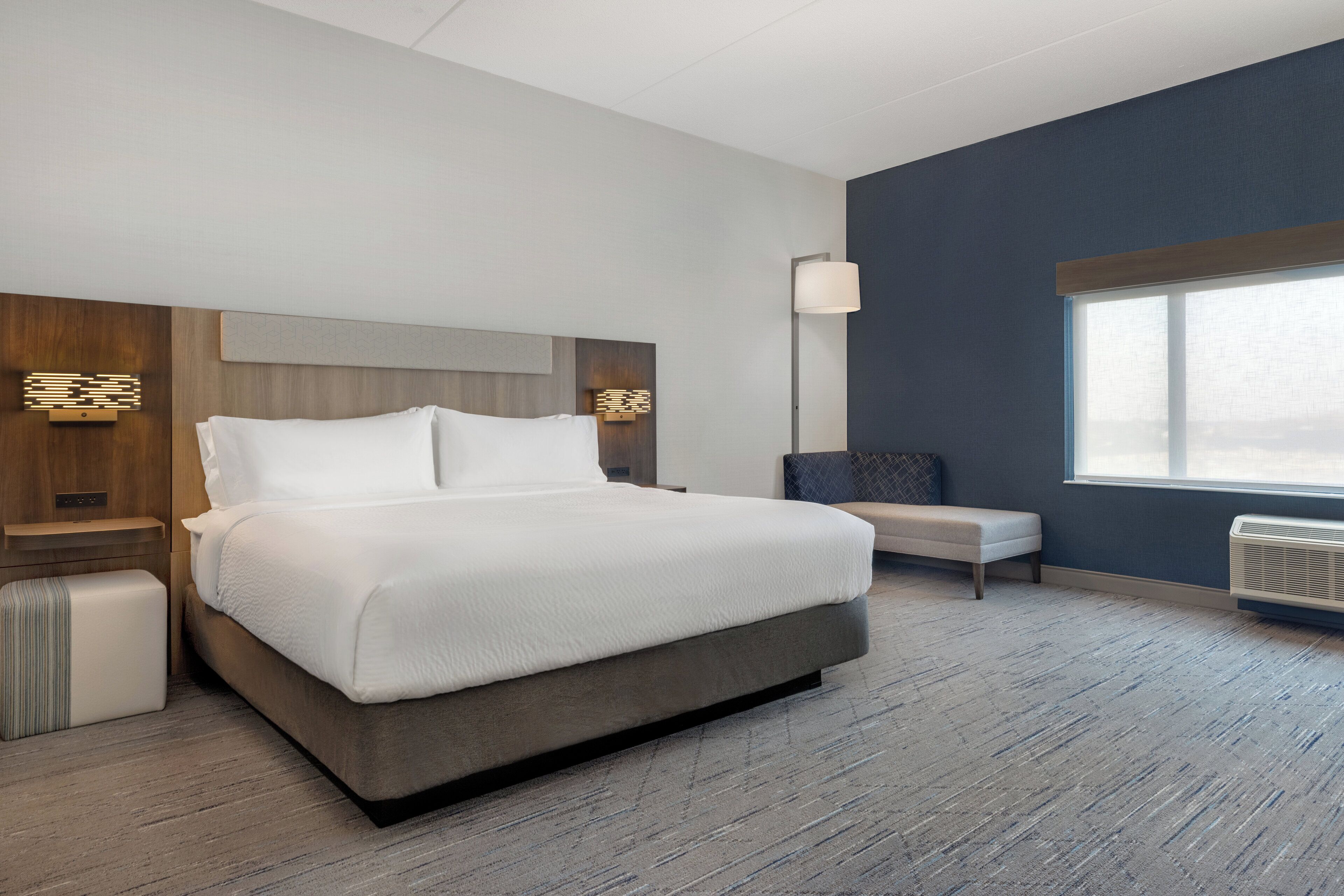 A modern hotel room with a large bed, a bench, a bedside table, and a window.