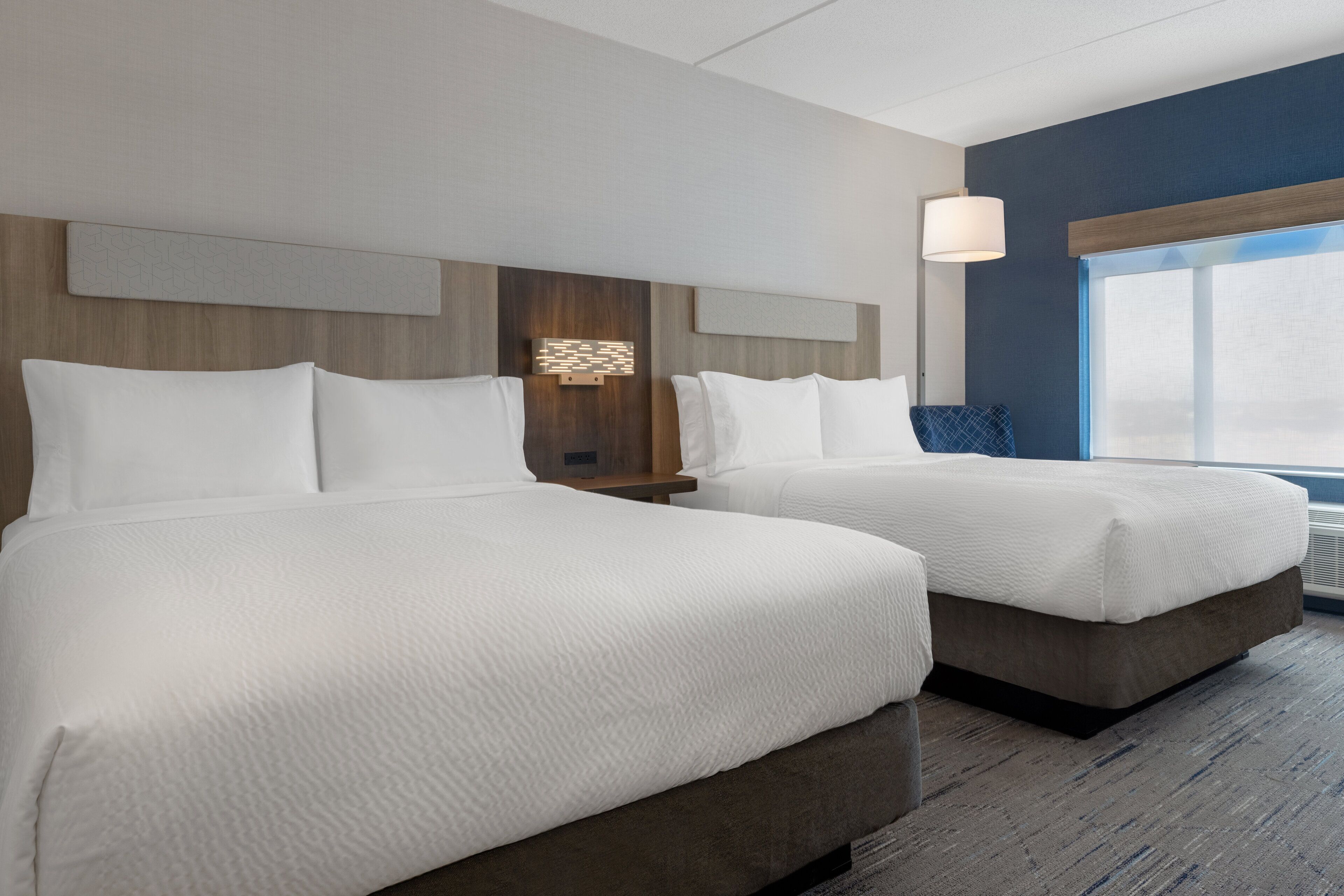 A hotel room with two beds, a wooden headboard, a wall-mounted light, and a large window.