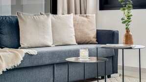 En moderne stue med en grå sofa, beige og hvide puder, et lille rundt bord med et glas og en bog, samt en vase med grønne planter.