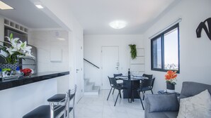 Une cuisine moderne avec un plan de travail noir, des armoires blanches et un coin repas comprenant une table et des chaises. Un escalier mène à un autre niveau, et il y a un canapé gris avec un coussin blanc.
