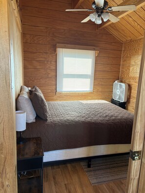 A wooden-paneled room with a bed, a ceiling fan, a window with blinds, and a portable heater.
