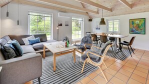 A spacious living area with a high ceiling, wooden beams, a sofa, chairs, a dining table, and large windows offering a view of greenery. - "Osmo" - 900m from the sea by Interhome (Ulfborg)