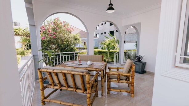 Un espacio de comedor al aire libre cubierto con una mesa y sillas de bambú, barandillas blancas y vistas a un jardín y un edificio.