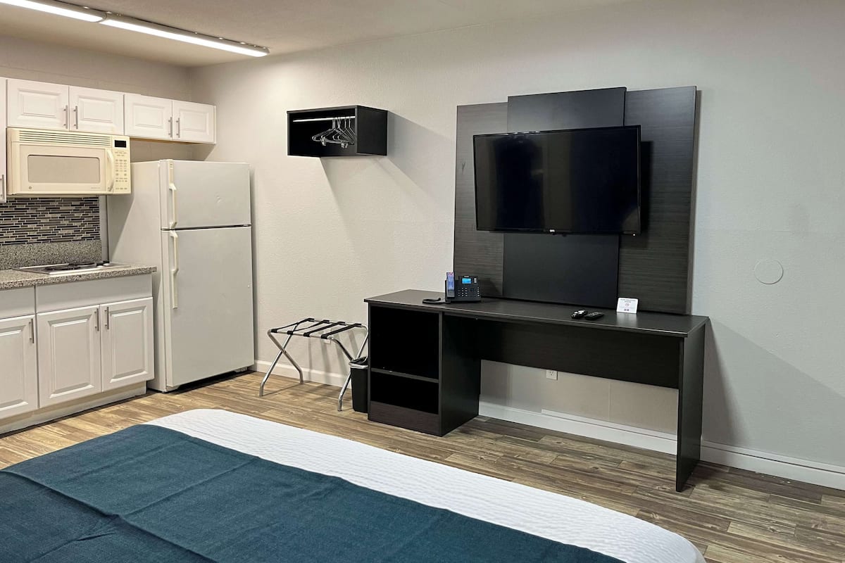 A compact hotel room with a bed, kitchenette, and a desk with a flat-screen TV.