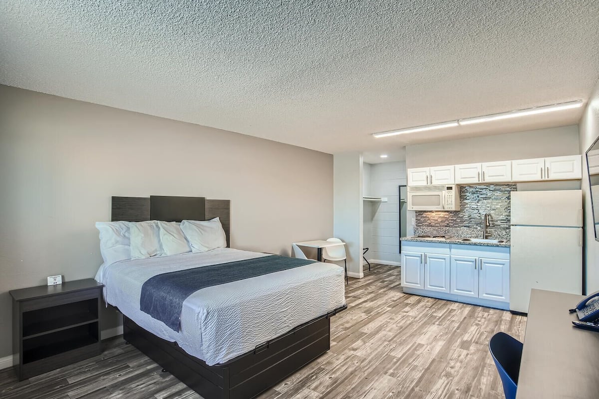 A bedroom with a bed, bedside tables, and a small desk with a chair. Adjacent is a kitchenette with a microwave, stove, and cabinets.