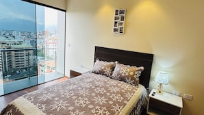 Un dormitorio moderno con una cama grande, una mesita de noche, una lámpara y vistas a la ciudad a través de ventanales de gran altura.